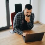 Young man with beard working on laptop at desk in a modern office setting.